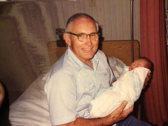 An elderly man smiles brightly while cradling a small baby in a warm, inviting room.