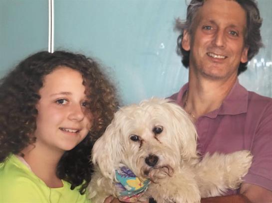 A girl and her father smile while holding their adorable white dog in a cozy living room.