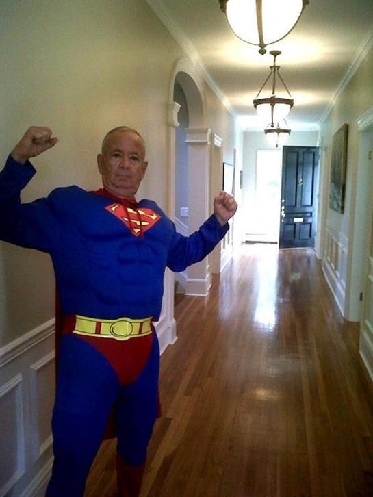 A senior man dressed as a superhero poses confidently in a spacious home corridor.
