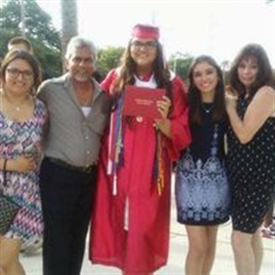 A student in a red graduation gown stands proudly with family members at the ceremony.