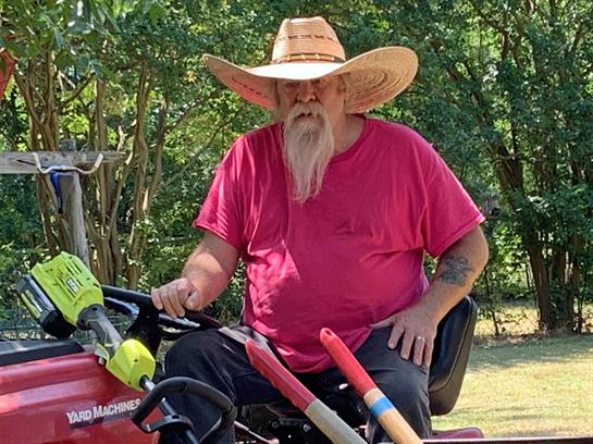 A man wearing a large hat sits on a red tractor, surrounded by greenery under bright sunlight.
