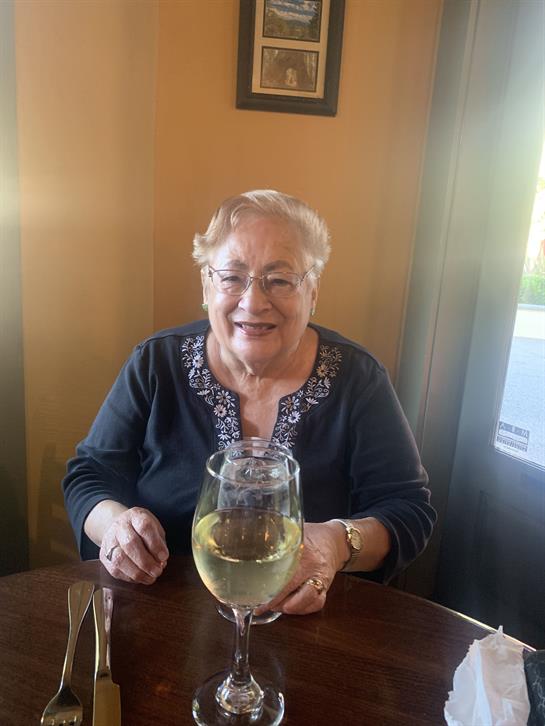 Older woman smiles while holding a glass of white wine at a small table in a restaurant.
