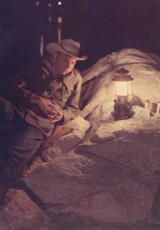 A cowboy relaxes by a lantern at a rocky campsite while dusk casts shadows around.