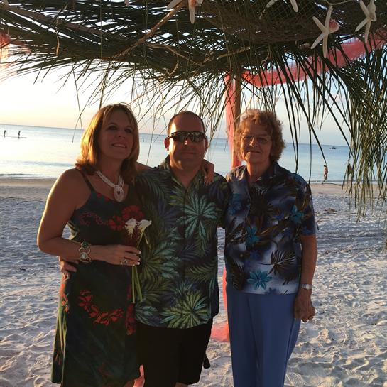 Three friends stand together on a sandy beach under palm branches, enjoying a beautiful sunset.