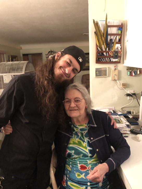 A young man smiles widely while embracing an elderly woman in a warm home environment.