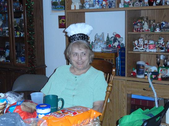 Senior woman wears a chef hat while seated at a table surrounded by family and snacks.