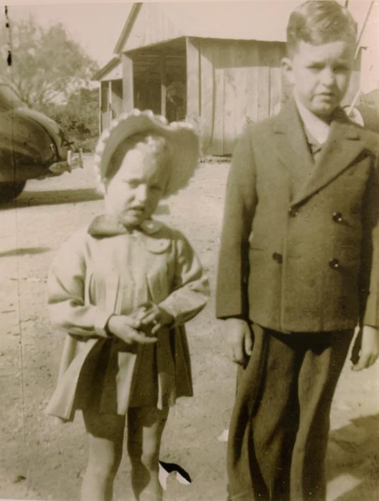 Two children pose outside a wooden structure, showcasing vintage fashion in a historic setting.