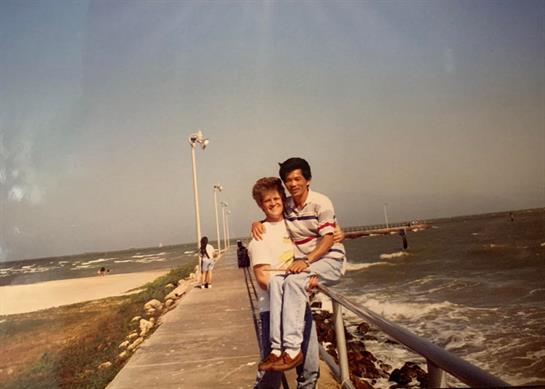 Two people share a joyful moment on a wooden pier overlooking the water with gentle waves.