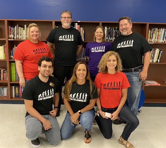 Group gathers in a school library wearing colorful shirts, smiling and engaging with each other.
