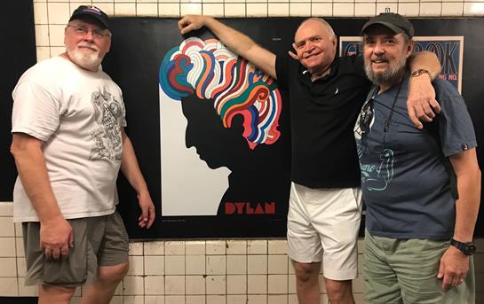 Three friends enjoy their time taking a photo by a vibrant mural showcasing Bob Dylan in a subway.