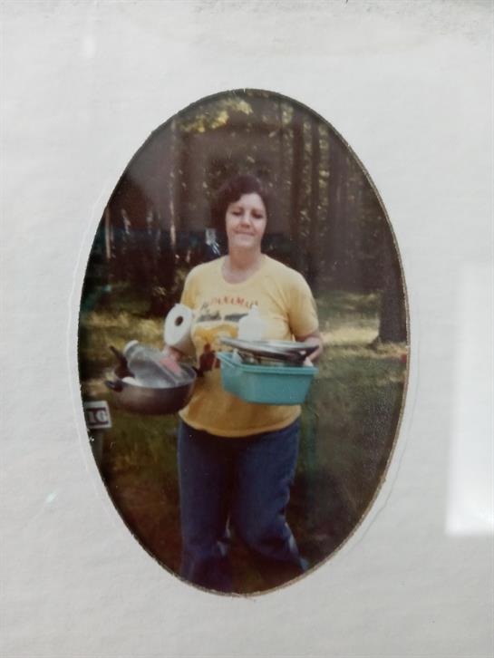 A woman balances various kitchen items while walking through a wooded area during the day.