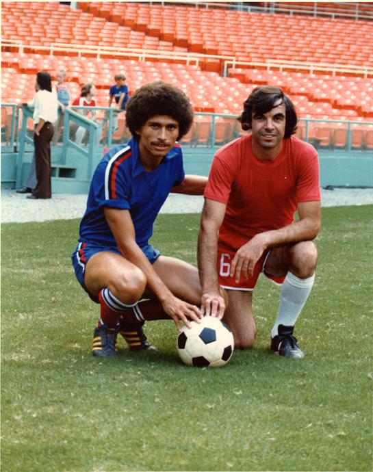 Two soccer players in colorful uniforms kneel together with a ball on the grass.