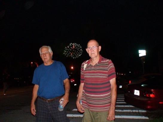 Two men stand together in a park, watching colorful fireworks light up the night sky.