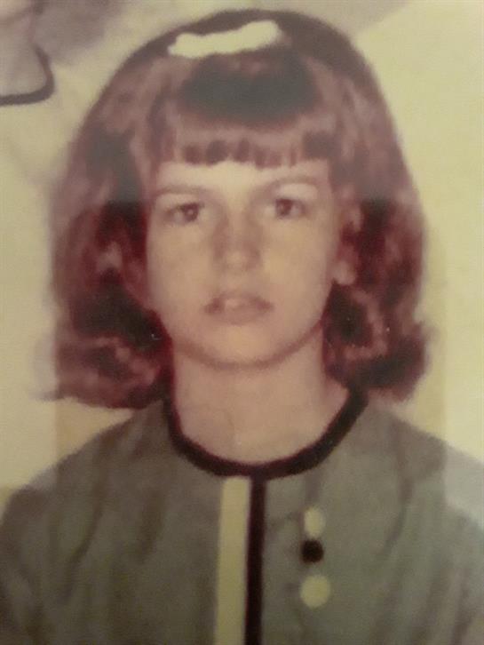 A young girl with wavy brown hair is posing in a green outfit against a simple background.