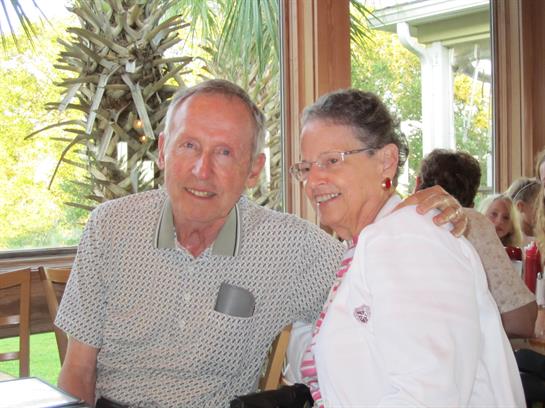 A cheerful elderly couple smiles warmly while seated at a café filled with sunlight and plants.