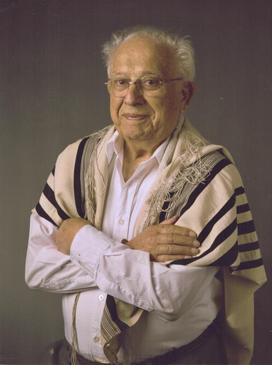 An elderly man in a tallit stands with arms crossed, symbolizing culture and spirituality.