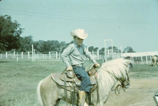 A young cowboy sits on a pony, enjoying a sunny day at the ranch surrounded by open fields.