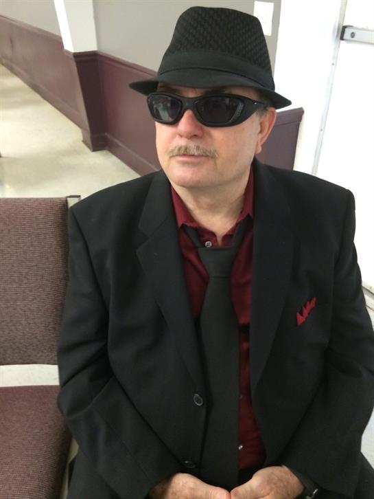 A man dressed in a black suit and hat sits quietly in a contemporary interior space.