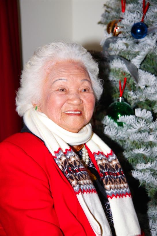 Smiling elderly woman in red attire poses cheerfully by a festive Christmas tree in winter.