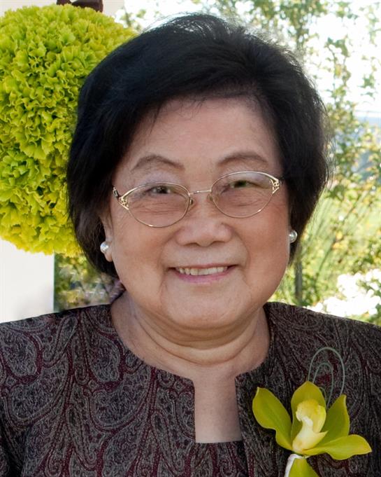 A woman wearing glasses and a flower accessory smiles happily in a vibrant garden setting.
