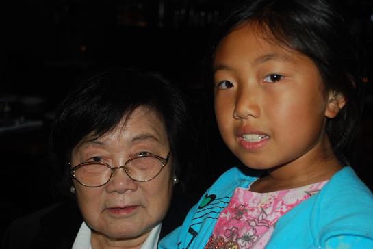 A grandmother and granddaughter share a warm moment during a family gathering under evening lights.