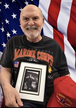 Elderly man displays a framed photo while standing against a backdrop of the American flag.