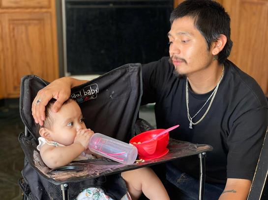 A father gently holds his baby's head while the little one drinks from a bottle in a high chair.