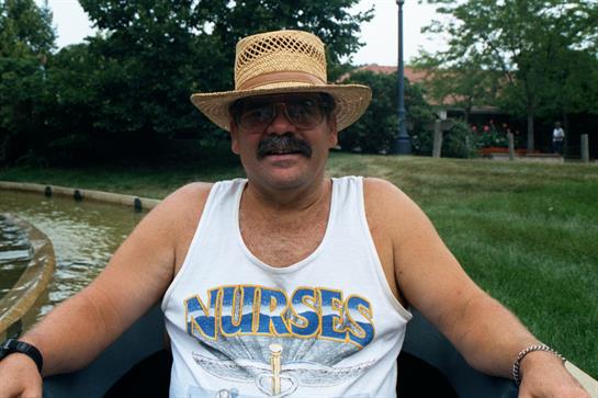 A man with a mustache wears a straw hat and tank top while relaxing in a park on a sunny day.