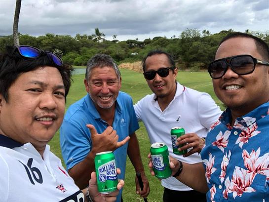 Four friends stand together on a golf course, celebrating with drinks and laughter on a sunny day.