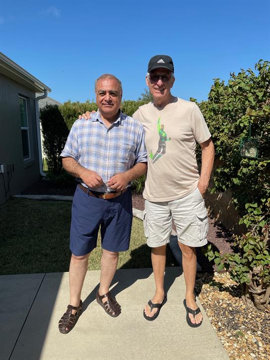 Two men stand together smiling in a well-kept garden on a bright sunny day.