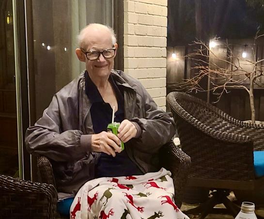 An elderly man sits comfortably on a porch with a warm blanket, sipping a drink on a calm evening.