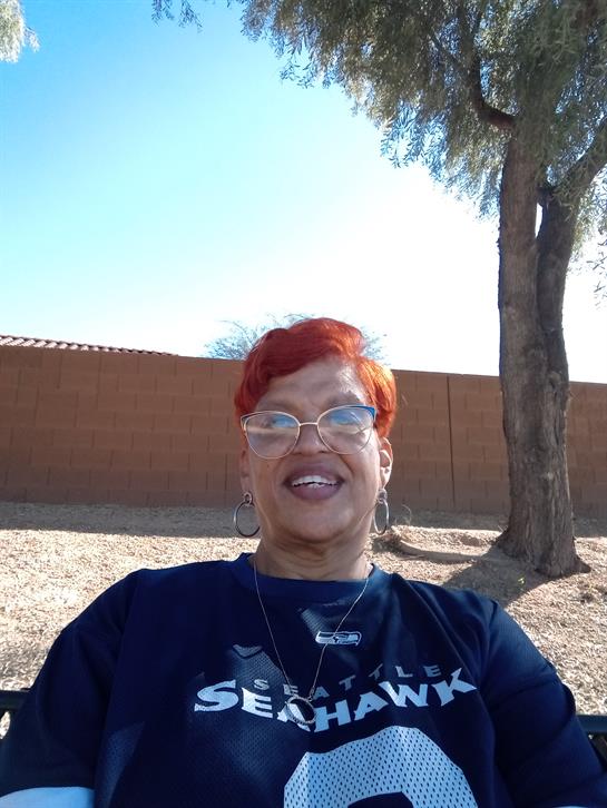 A cheerful person with bright red hair relaxes under a tree in a park, enjoying the sunny weather.