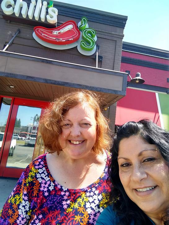 Two women smile happily outside a lively restaurant on a sunny day.