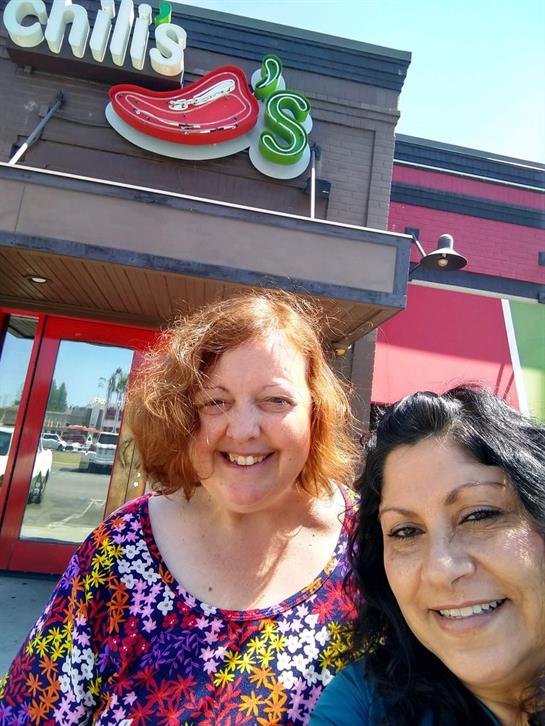 Two happy friends pose for a selfie in front of a colorful restaurant on a bright day.