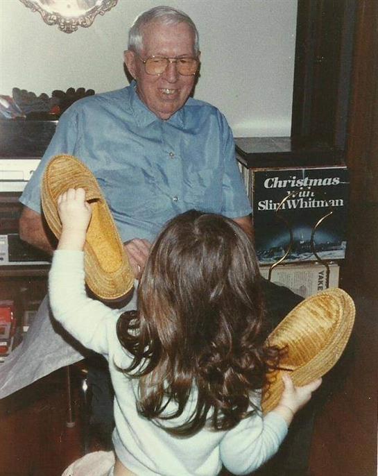 An elderly man enjoys a light moment as a young girl playfully presents him with slippers at home.
