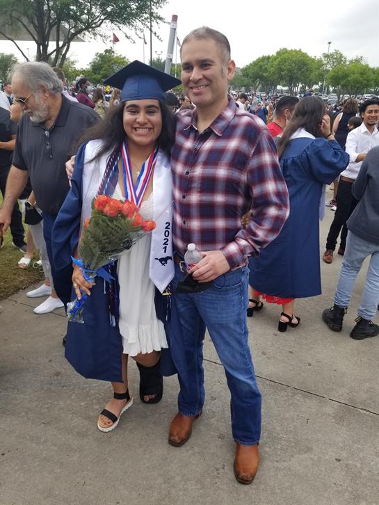 A graduate smiles while holding a bouquet and posing with a family member at a graduation event.