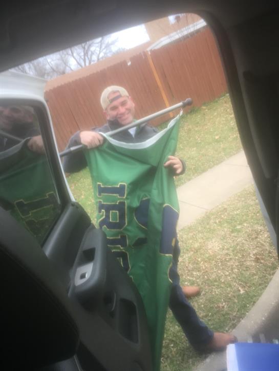 A man displays a large green banner while standing by a parked vehicle in a grassy area.