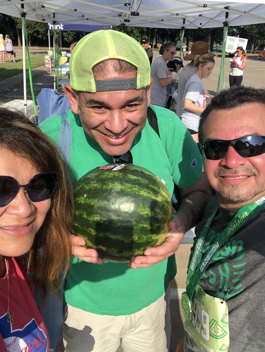 Three friends smile while holding a watermelon at a summer festival in a vibrant outdoor setting.