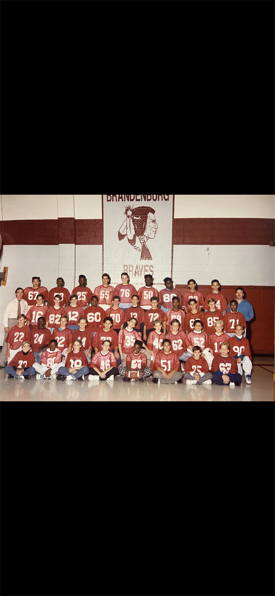 Student athletes gather in the gym wearing red football jerseys with numbers for a team photo.