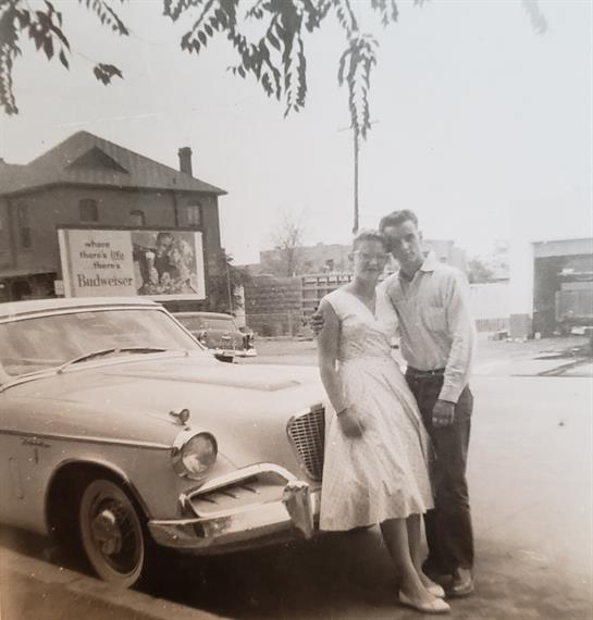 A couple stands together smiling near a vintage car in a quaint urban setting.