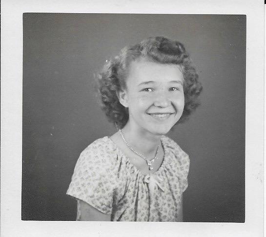 A smiling young girl with curly hair poses in a vintage setting, wearing a light-patterned dress.
