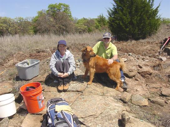 Two volunteers engage in conservation efforts outdoors, accompanied by a dog at a natural landscape.