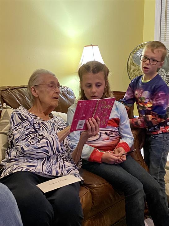 An elderly woman shares a storybook with two engaged children in a warm living room.