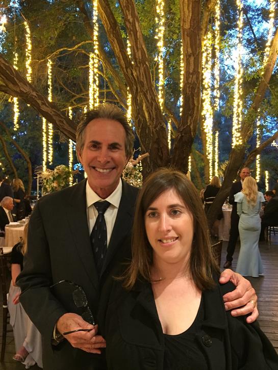 A couple poses happily amidst twinkling string lights and guests at a festive outdoor gathering.