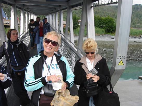 Two tourists smile as they stroll along a boarding bridge in Alaska on a sunny day.