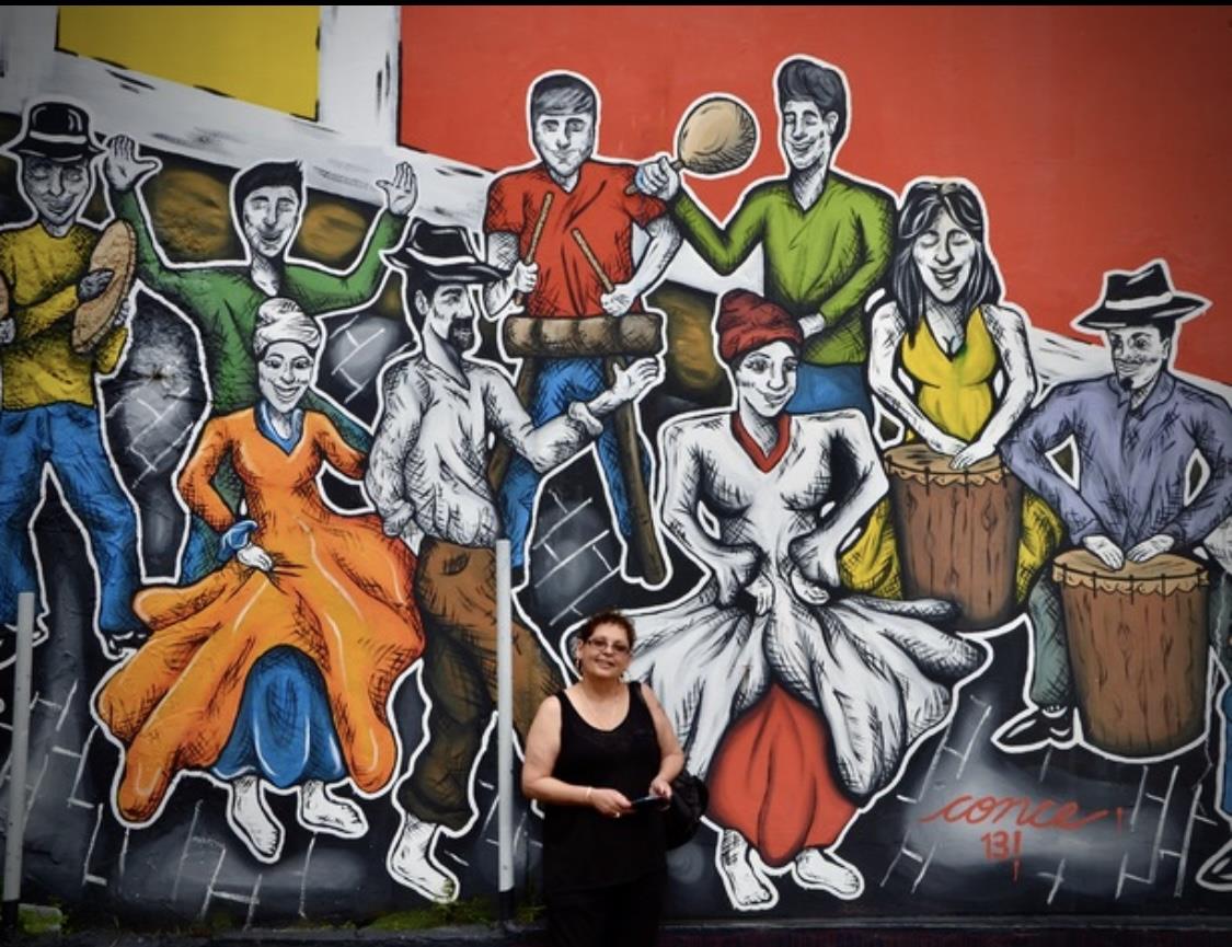 A woman stands smiling in front of a vibrant mural showcasing dancers in colorful attire.