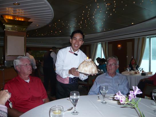 A waiter in formal attire serves dessert to two older men at a white tablecloth dinner.