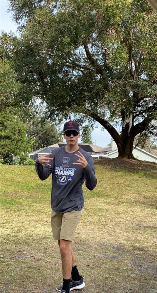 A man confidently poses in a park, wearing sunglasses and a casual outfit under a clear sky.