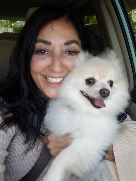 A woman smiles brightly while holding her fluffy white dog in a vehicle, showcasing their bond.