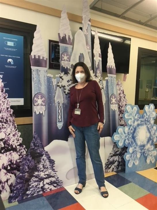 A woman stands next to a winter-themed backdrop with snowflakes and trees indoors.
